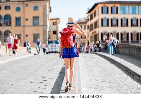 Beautiful female traveler in blue dress, hat and backpack walking on crowded Holy Trinity bridge in Florence city. Back view