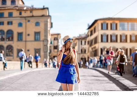 Beautiful female traveler in blue dress, hat and backpack walking on crowded Holy Trinity bridge in Florence city. Vacation in Italy