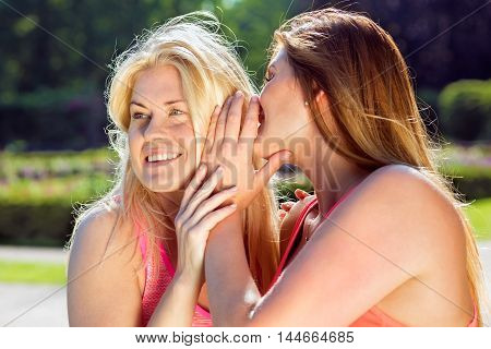 Pair of smiling blond and brunette women sharing secrets or gossip news with each other outside