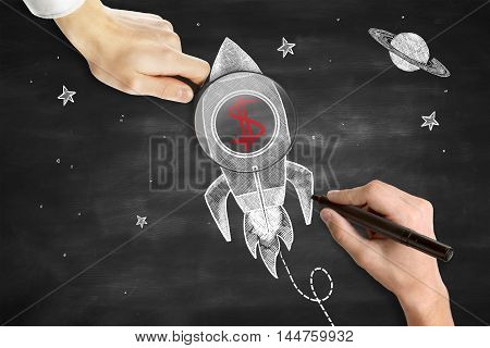Male hands drawing and holding magnifier over rocket ship sketh with red dollar sign. Blackboard background. Start up concept