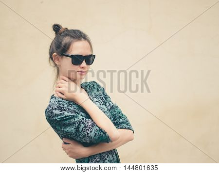 Fashionable serious young woman in black sunglasses posing near wall with copy space