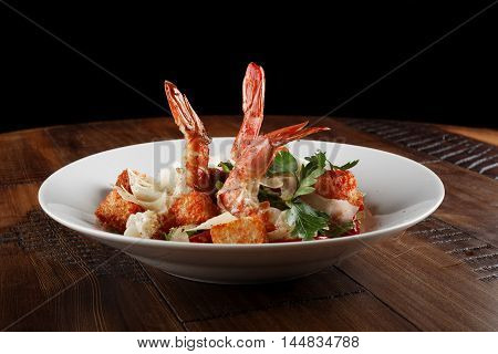 Caesar salad with king prawns in a white plate. Salad with shrimp on wooden brown background.