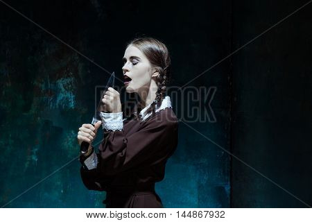 Portrait of a young girl with knife in school uniform as killer woman against school board . The image in the style of Halloween and Addams family