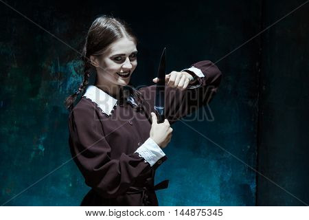 Portrait of a young girl with knife in school uniform as killer woman against school board . The image in the style of Halloween and Addams family