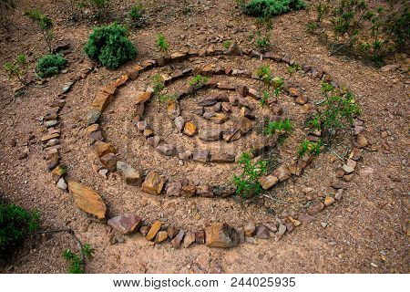 Stone Spiral Made Up Of Lots Of Individual Rocks On A Dry Stony Floor With Green Plants Growing Up T