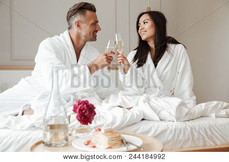 Happy lovely couple caucasian man and asian woman wearing white housecoat smiling to each other and drinking sparkling wine during breakfast in bed at hotel room