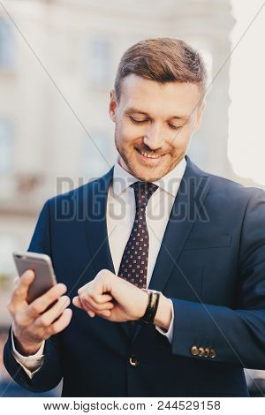 Photo Of Clever Male Lawyer Waits For Client Outdoor, Looks At Wristwatch, Holds Smart Phone As Wait