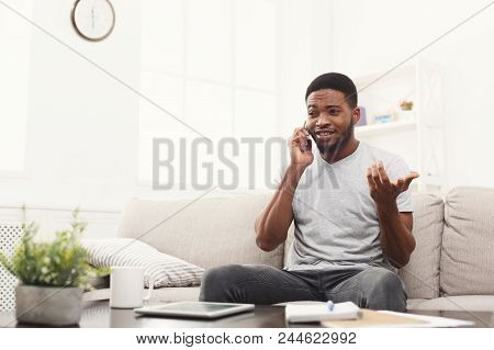 Happy Young African-american Man Talking Emotionaly On Smartphone Sitting On Beige Couch At Home, Co
