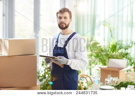 Serious confident bearded young male mover in overall and gloves holding sketchpad and looking at camera while checking delivered goods in new restaurant