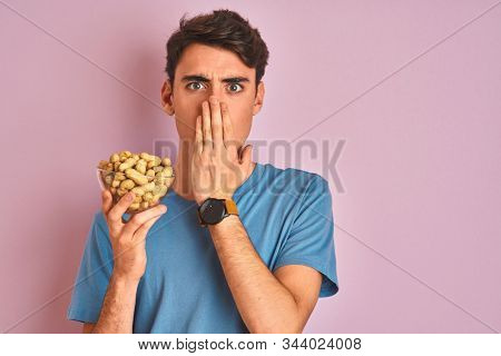 Teenager boy holding a bunch of peanunts over isolated pink background cover mouth with hand shocked with shame for mistake, expression of fear, scared in silence, secret concept
