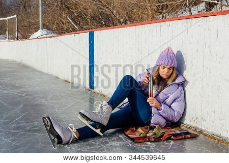 Lovely Young Woman Relaxing After Riding Ice Skates And Drinking Hot Drink From Termo Pot On The Ice