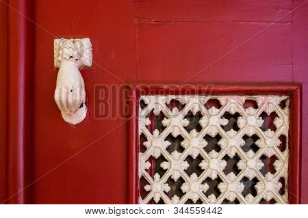 Bright Red Painted Wooden Entrance Door With A White Knocker In A Shape Of A Hand And An Ornamental 