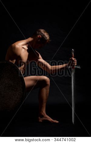 Side View Of Young Strong Warrior Standing On Knee And Looking Down. Muscular Half Naked Athlete Pos