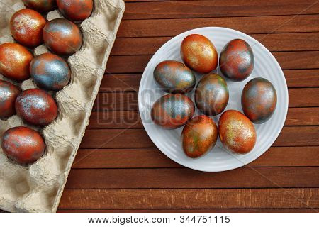 Brown, Gold And Blue Painted Easter Eggs Lie In A White Plate And On A Stand On A Brown Wooden Table