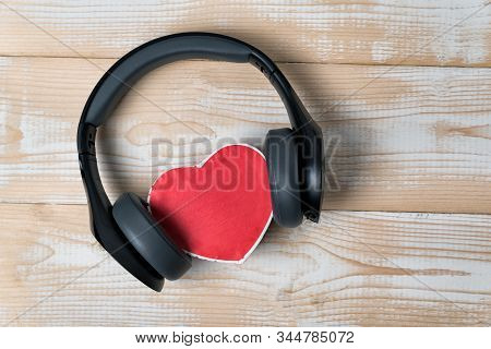 Full-size Wireless Headphones Pulled Over Small Red Heart-shaped Box On A Light Brown Wooden Table. 