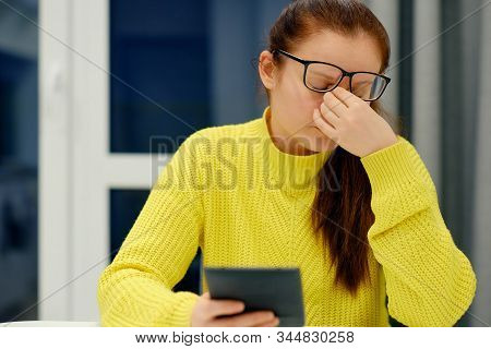 Young Caucasian Female In Glasses And Knitted Sweater Doing The Nose Massage When Reading Electronic