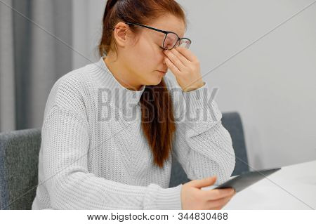 Young Caucasian Female In Glasses And Knitted Sweater Doing The Nose Massage When Reading Electronic
