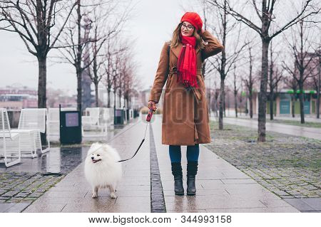 Beautiful Smiling Girl In A Red Beret And A Brown Coat Walks With A White Pomeranian Spitz In Autumn