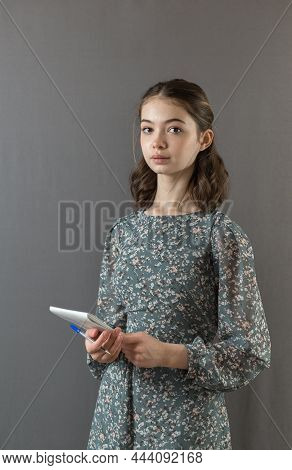 A Beautiful Serious Successful Girl In A Dress Stands Against A Gray Wall With A Notebook And Pen In