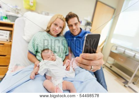 Man with woman and newborn babygirl taking selfportrait through cell phone in hospital room
