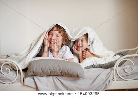 Two children brother and sister squirmy on the bed in the bedroom. They with a smile look out from under a blanket.