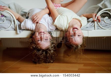 The children boy and girl naughty on the bed in the bedroom. They lie side by side their heads hanging off the bed. Children is very amused.