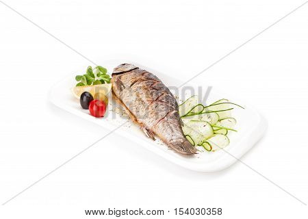 Isolated delicious - the freshly cooked, fried fish - crucian served with a cucumber, an olive and a lemon on a white dish.