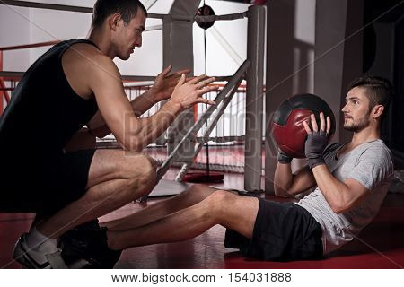 Throwing back. Young handsome bearded man training abdomen while throwing ball to with his trainer.