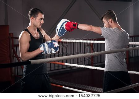 Direct hook. Side view of young bearded man standing on ring and working on boxing hook together with his trainer in special pad.