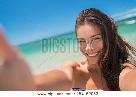Happy joyful asian young woman enjoying summer holidays taking selfie phone picture swimming in the ocean water on beach vacation. Cute chinese girl smiling at camera taking a swim self-portrait.