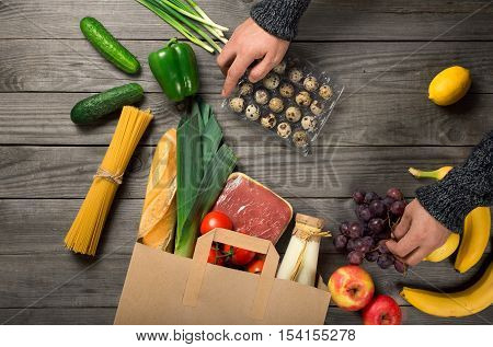 Man looks brown paper bag full of different healthy food on wooden kitchen table top view
