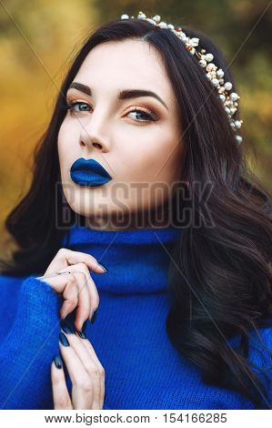 Portrait of beautiful young woman with blue lips and blue sweater with accessory on her head in the park