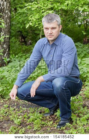 Portrait of grey-haired man in checkered shirt sits squatted at summer park.