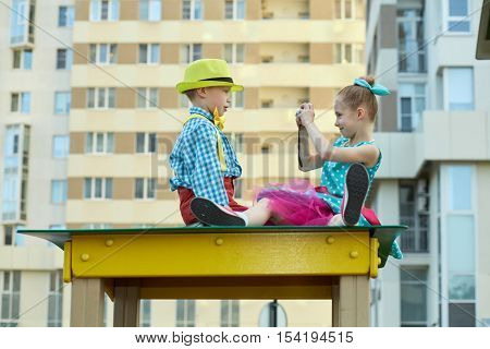 Little girl takes picture of little boy sitting on roof of wooden construction at playground.