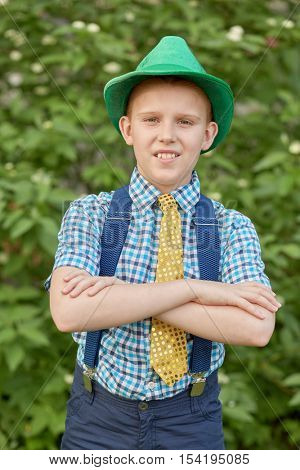 Boy in hat with arms folded on chest against green bushes.