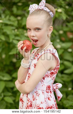 Girl in flowery gown going to bite red apple standing outdoor against green bushes.
