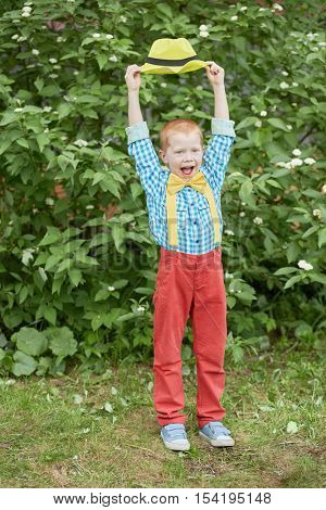 Little red-haired boy with open mouth dressed in dance suit holds yellow hat above his head outdoors.