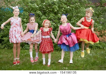 Five smiling little girls dressed in dancing suits pose jumping up at grassy lawn.