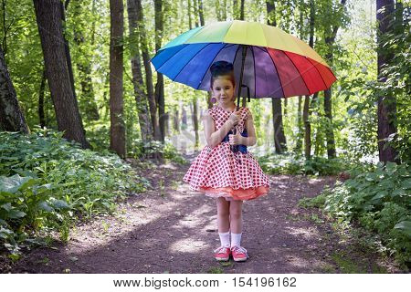 Little girl in polka-dotted dress stands with rainbow coloured umbrella in summer park.