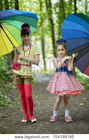 Two girls stands with rainbow coloured umbrellas in summer park.