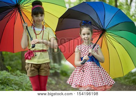 Two girls stand with rainbow coloured umbrellas in summer park.