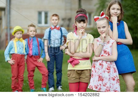 Three girls propping chins at foreground and three boys behind them out of focus stand outdoor on summer day.