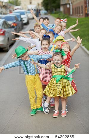 Fifteen children stand one by one outstretching arms to sides at sidewalk, focus on first girl.