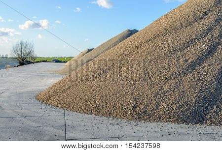 Large heaps of sand and gravel stored on concrete slabs next to a wide Dutch river. It is a sunny day in the middel of the winter season.