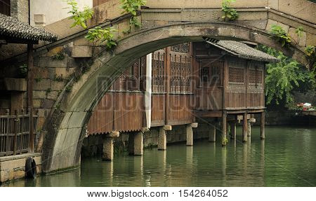 A wooden building lining the water canals as seen under a weathered curved stone bridge within Tongxiang's Wuzhen Scenic Area in Zhejiang province China.