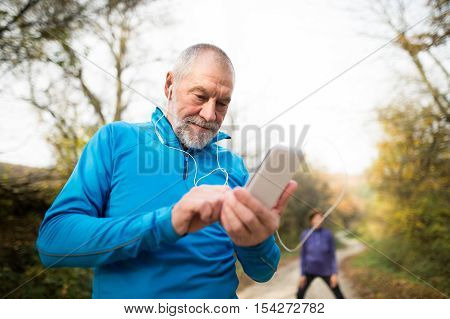 Senior runners in nature, stretching. Man with smart phone with earphones. Listening music or using a fitness app. Using phone app for tracking weight loss progress, running goal or summary of his run.
