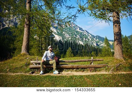 Wide shot of bearded man who is looking at the sky while sitting on the bench. Enjoying stunning vista of mountains on a cloudless gorgeous day 