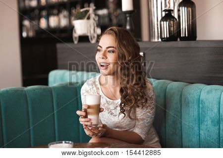 Laughing girl 20-24 year old sitting in cafe drinking coffee/ Wearing stylish white dress.