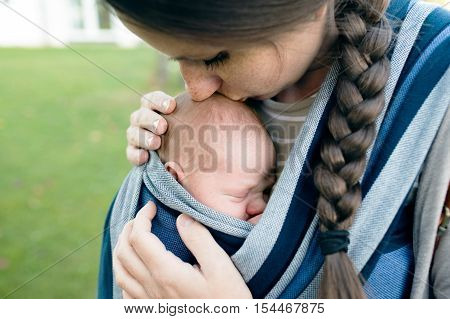 Beautiful young mother kissing her newborn baby son in sling outside in green nature.