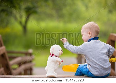 Cute little toddler boy sitting on wooden bridge and playing with white teddy bear in the park on sunny day. Happy child playing with toy. Children playing outdoors. Lifestyle and childhood concept